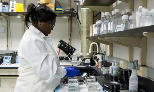 A woman in a lab, wearing a white lab coat and gloves. 